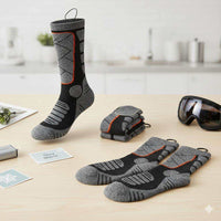 Gray and black rechargeable heated socks with orange accents displayed on a table with ski goggles and winter-themed cards